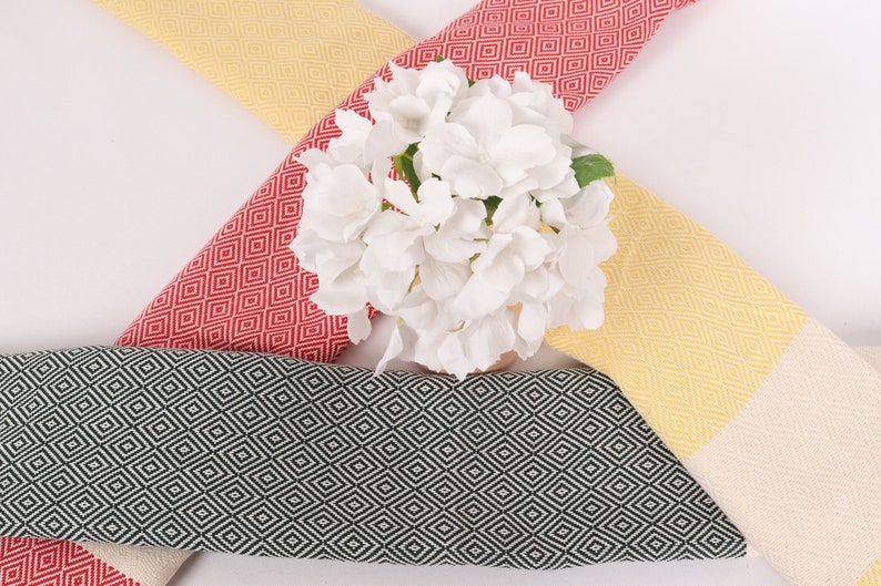 May include: Three Turkish towels with a diamond pattern in yellow, red and black. The towels are arranged in a cross shape with a bouquet of white flowers in the centre.