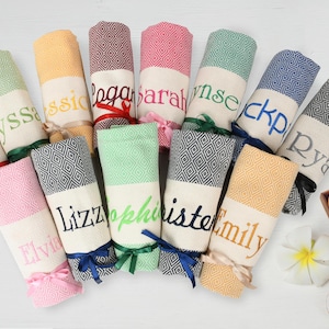 May include: A collection of 11 personalised towels with various colours and names embroidered on them. The towels are folded and tied with ribbons.