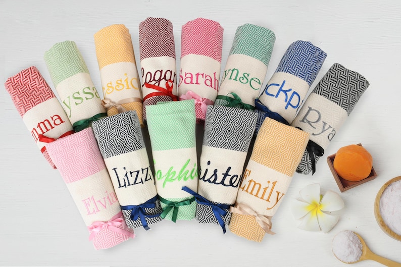 May include: A collection of 11 personalised towels with various colours and names embroidered on them. The towels are folded and tied with ribbons.