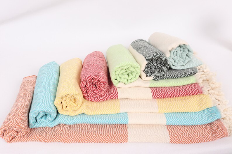 May include: A stack of colourful Turkish towels with fringe, folded and rolled, in shades of orange, yellow, blue, green, and red.