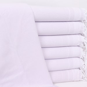May include: A stack of six white Turkish towels with fringed edges. The towels are neatly folded, showcasing a clean, minimalist aesthetic. A vase with white flowers is visible in the background, adding a touch of elegance.