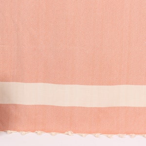 May include: A peach-coloured Turkish towel with a diamond pattern and a white stripe. The towel has tassels along the bottom edge.