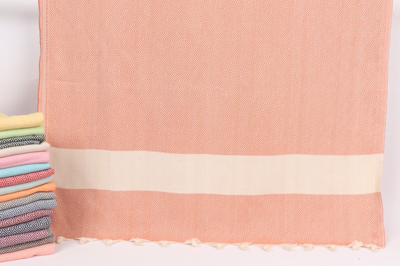 May include: A peach-coloured Turkish towel with a diamond pattern and a white stripe. The towel has tassels along the bottom edge.