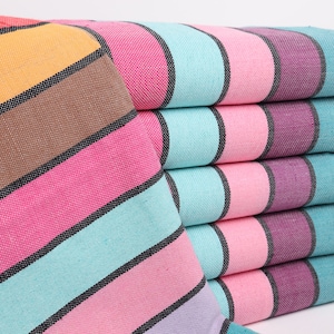 Puede incluir: Una pila de toallas rayadas de colores con un borde con flecos. Las toallas están dobladas y dispuestas en un patrón de arcoíris, con colores que incluyen rojo, naranja, amarillo, marrón, rosa, turquesa y morado.