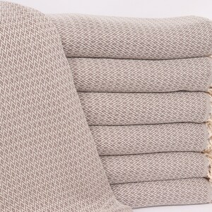 May include: A stack of six beige cotton throw blankets with a diamond pattern and fringe. The blankets are folded and stacked on a white surface.