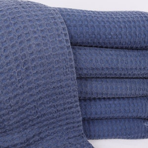 May include: A stack of six blue waffle weave towels with fringed edges. The towels are folded and stacked on a white surface.