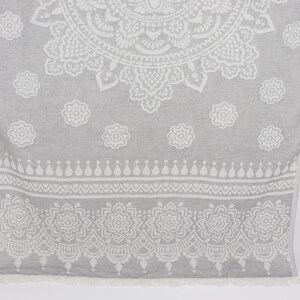 May include: A gray and white patterned Turkish towel with a fringe border. The towel features a large mandala design in the center and smaller floral patterns around the edges.
