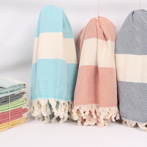 May include: Three Turkish towels, one blue, one peach, and one grey, with white stripes and diamond patterns. The towels are hanging from a white background.