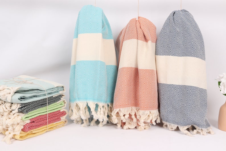 May include: Three Turkish towels, one blue, one peach, and one grey, with white stripes and diamond patterns. The towels are hanging from a white background.