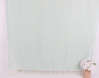 May include: A light green Turkish towel with white fringe. The towel is hanging on a white wall with a white flower arrangement in a vase on the bottom right corner.