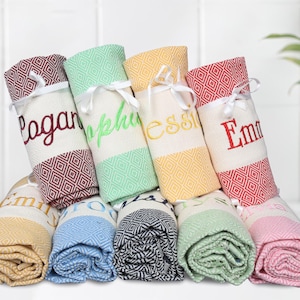May include: Eight personalized towels rolled up and tied with white ribbon. The towels are in various colors, including red, green, yellow, blue, black, and pink. The names Logan, Sophia, Essie, Emma, Emily, Owen, and Ava are embroidered on the towels.