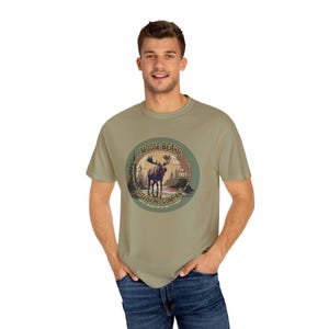 May include: A khaki green t-shirt with a vintage-style graphic of a moose in a forest setting. The graphic includes the text "Moose Beard Provisions Company Est. 1907".