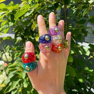 Puede incluir: Un conjunto de seis anillos de resina coloridos con detalles dorados. Cada anillo presenta un color diferente y un patrón único. Los anillos están apilados en una mano.