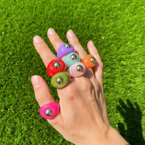 Puede incluir: Un maniquí de mano con un conjunto de siete anillos coloridos con detalles dorados. Los anillos están hechos de resina y tienen una variedad de texturas y patrones.
