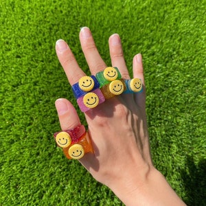 Pode incluir: Uma mão usando vários anéis coloridos com carinhas sorridentes. Os anéis são feitos de resina e têm glitter embutido.