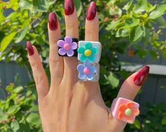 May include: Five square rings with flower designs are shown. The rings have a black, white, clear, and pink base, with flower colours of purple, green, blue, and orange. The background is a blurred green, with the rings on a hand.