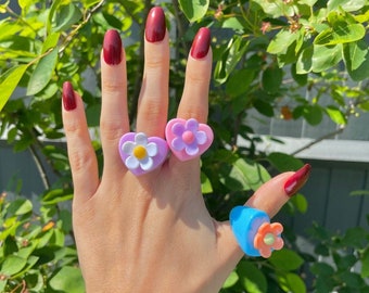 May include: Three colourful flower-shaped rings. The rings are made of plastic and feature a heart-shaped base with a flower design. The rings are pink, purple, and blue.