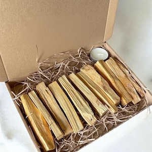 Op de afbeelding: Een open kartonnen doos met verschillende Palo Santo-stokken en een kleine witte kaars. De Palo Santo-stokken zijn lichtbruin en gerangschikt met decoratief stro.