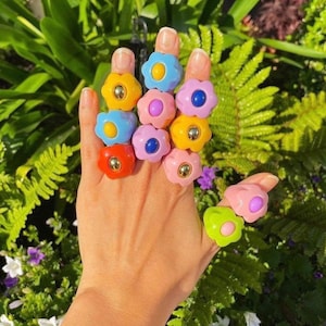 May include: A collection of colorful flower-shaped rings in various shades of yellow, blue, pink, red, and green. Each ring features a gold-colored center. The rings are displayed on a hand against a backdrop of green foliage.