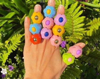 May include: A selection of flower-shaped rings in shades of yellow, blue, pink, red, and green. Each ring has a gold-coloured centre. The rings are displayed on a hand against a backdrop of green foliage.
