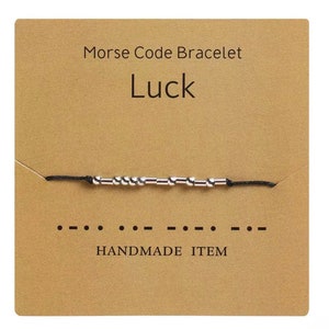 May include: A black string bracelet with silver beads spelling out the word "Luck" in Morse code. The bracelet is displayed on a brown card with the text "Morse Code Bracelet" and "Handmade Item".