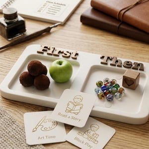 May include: A white rectangular tray with two recessed sections labeled First and Then in dark wooden letters. The tray contains three dark brown round treats, a green apple, a small wooden cube with a heart, and several colorful glass beads.