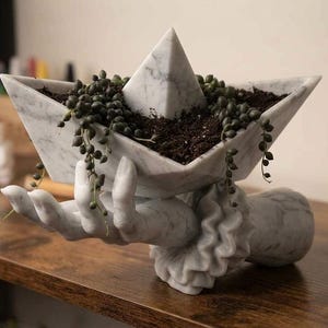 May include: A white marble-effect planter shaped like a paper boat, held by a sculpted hand with a ruffled cuff. The boat contains dark soil and a trailing string of pearls plant with small green spherical leaves, creating a decorative display.