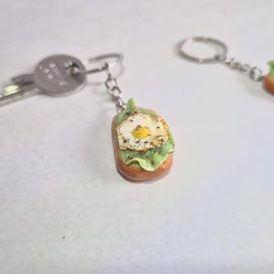 Könnte beinhalten: Schlüsselanhänger mit Miniatur-Lebensmittelrepliken. Ein Schlüsselanhänger zeigt einen Schlüssel neben einem Miniatur-Sandwich mit Spiegelei. Der andere Schlüsselanhänger zeigt ein Miniatur-Sandwich mit Spiegelei. Die Schlüsselanhänger befinden sich auf einer weißen Oberfläche.