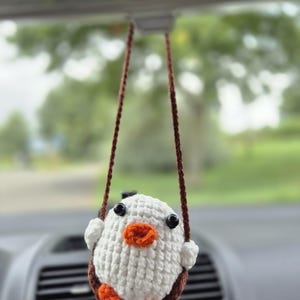 Könnte beinhalten: Ein handgefertigter, weißer Häkelvogel-Autoanhänger mit schwarzen Augen und einem orangefarbenen Schnabel und Füßen. Der Vogel wird von braunen Kordeln gehalten, die am Rückspiegel eines Autos hängen. Der Anhänger ist ein skurriles Accessoire.