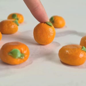 Könnte beinhalten: Eine Sammlung kleiner, orangefarbener Marzipan-Orangen mit grünen Blättern. Die Orangen sind auf einer weißen Oberfläche angeordnet.