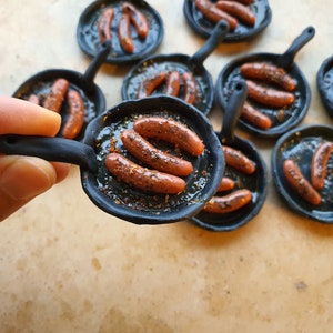Könnte beinhalten: Ein Set aus Miniatur-Gusseisenpfannen mit schwarzen Griffen, jede gefüllt mit kleinen, braunen Würstchen. Die Pfannen sind auf einer hellbraunen Oberfläche angeordnet.