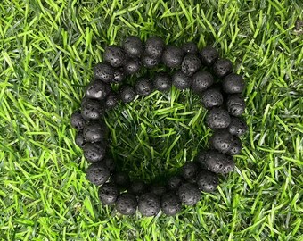 Puede incluir: Un brazalete de cuentas de roca de lava negra. El brazalete está hecho de pequeñas cuentas redondas con una superficie rugosa y texturizada. Las cuentas están enhebradas en un cordón negro.