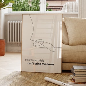 May include: A framed print with a minimalist line drawing and the text "existential crisis can't bring me down". The print is hanging on a white wall in a living room with a potted plant, a wooden chair, and a couch.