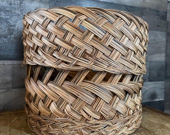 May include: A cylindrical woven basket made from natural fibres. The basket has a lid and is constructed with a crisscross weave pattern. The colour is a natural, light brown, and the overall style is rustic and handmade.