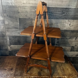 May include: A two-tiered wooden serving stand featuring a medium brown finish. The structure consists of two rectangular shelves with scalloped edges, supported by four vertical legs that converge at the top into a single handle for carrying.