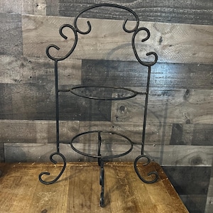 Könnte beinhalten: Schwarzer, schmiedeeiserner, mehrstöckiger Ständer mit dekorativen Schnörkeln. Der Ständer hat drei ovale Plattformen und einen bogenförmigen oberen Teil, der sich zum Ausstellen von Gegenständen eignet. Der Ständer ist etwa 61 cm hoch.
