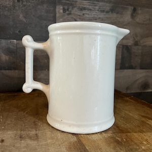 May include: A white ceramic pitcher with a handle and a spout. The pitcher has a cylindrical body and a decorative handle. The pitcher is sitting on a wooden surface.