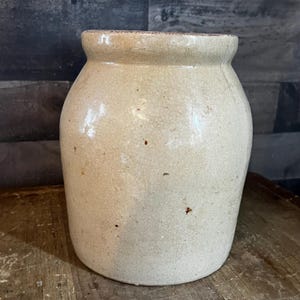 Peut inclure: Un pot en grès vintage de couleur blanc cassé, avec une large ouverture et un corps légèrement conique. La surface présente une texture craquelée et de petites taches brunes. Le pot est posé sur une surface en bois.