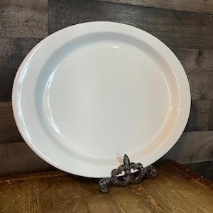 Może przedstawiać: Duży, okrągły, biały ceramiczny półmisek do serwowania. Półmisek jest podparty ozdobnym, bogato zdobionym metalowym stojakiem. Tło to ciemna drewniana powierzchnia.