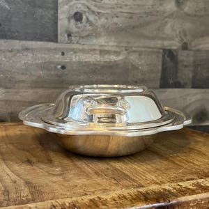 May include: A silver-colored, lidded serving dish with a scalloped edge and two handles. The dish is round with a slightly flared base and sits on a wooden surface. The background is a gray, textured wall.
