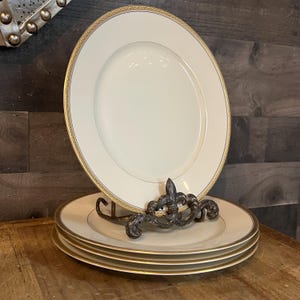 Puede incluir: Una pila de platos de porcelana blanca con un borde dorado y estampado. Los platos se exhiben en un soporte de metal decorativo con un diseño de flor de lis. Los platos están sobre una superficie de madera.