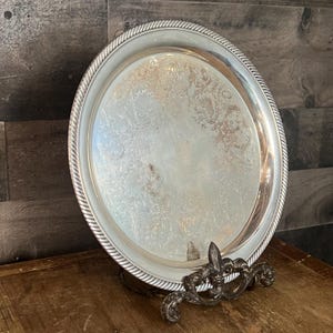 May include: A silver-colored, oval serving tray with a decorative rope-like border. The tray has an ornate floral pattern in the center and is supported by a decorative metal stand. The tray is displayed on a wooden surface.