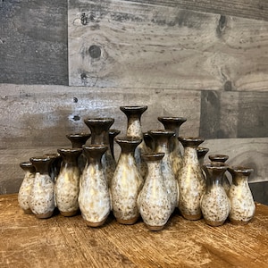 May include: A collection of 15 small, speckled ceramic vases in various sizes and shapes. The vases are arranged in a pyramid shape and are all a light brown color with white speckles.