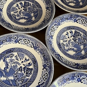 May include: A collection of white and blue ceramic plates with a classic Willow pattern. The plates feature a central landscape scene with birds and buildings, surrounded by a detailed border. The plates are arranged on a dark surface.