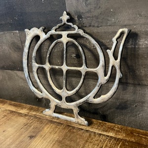 May include: Silver teapot-shaped trivet with a circular design. The trivet is made of metal and has a decorative, ornate design.