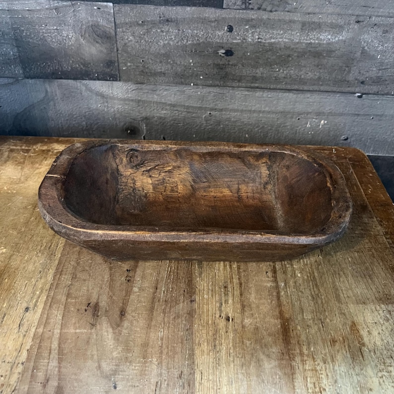 May include: A rustic wooden dough bowl with a natural brown finish. The bowl is oval shaped and has a slightly raised rim.