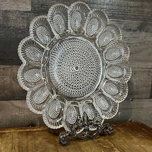 Peut inclure: Plat à œufs en verre transparent avec un bord festonné et des compartiments individuels pour les œufs. Le centre du plat présente un motif texturé et pointillé. Le plat est présenté sur un support en métal décoratif.