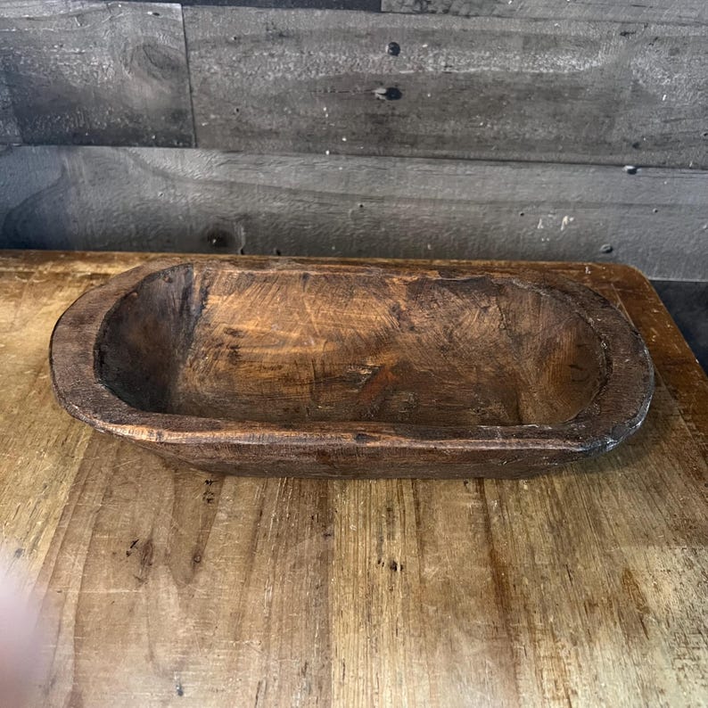 May include: A rustic wooden dough bowl with a natural brown finish. The bowl is oval shaped and has a slightly raised rim.