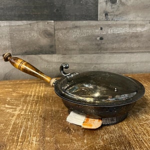 May include: A silver-plated, round, covered dish with a wooden handle. The dish has a beaded edge and a fluted base. The handle is brown and has a decorative knob.