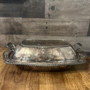 May include: A silver-colored, rectangular serving dish with a lid. The dish has a decorative rope-like trim around the edges and handles on either side. The dish sits on a wooden surface.